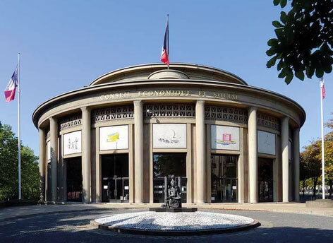 Bâtiment circulaire à colonnes, drapeaux français de part et d'autres, rond point en premier plan et ciel bleu