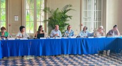 Dans un intérieur de bâtiment à hautes fenêtres et partère de carrelage, huit personnes sont alignées assis à une table au drapé bleu.