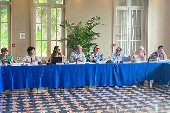 Dans un intérieur de bâtiment à hautes fenêtres et partère de carrelage, huit personnes sont alignées assis à une table au drapé bleu.