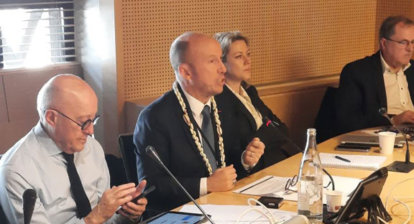 Le président parle assi à une table de réunion, un homme en premier plan regarde son téléphone