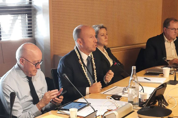 Le président parle assi à une table de réunion, un homme en premier plan regarde son téléphone