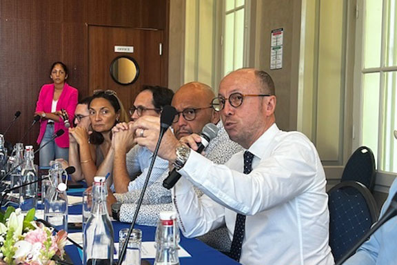 Un homme parle dans un micro, il est assis à une table de type tribune dans une salle à haut plafnod.